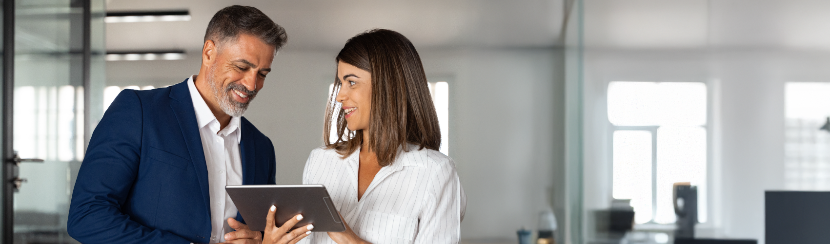 A man and a woman in office attire, looking at a tablet in the possession of the woman.