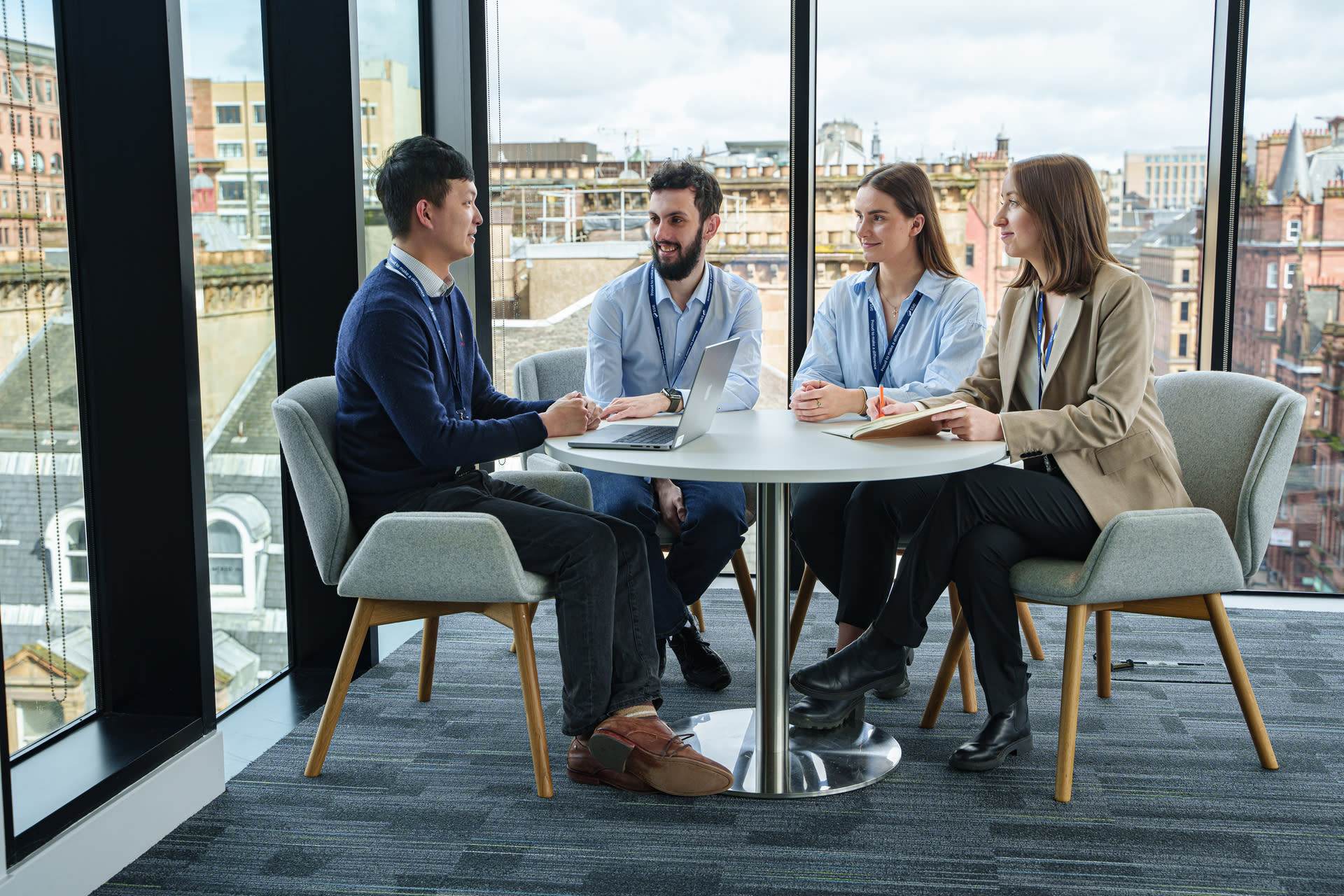 A group of people in office clothes sat around a desk with a city skyline in the background