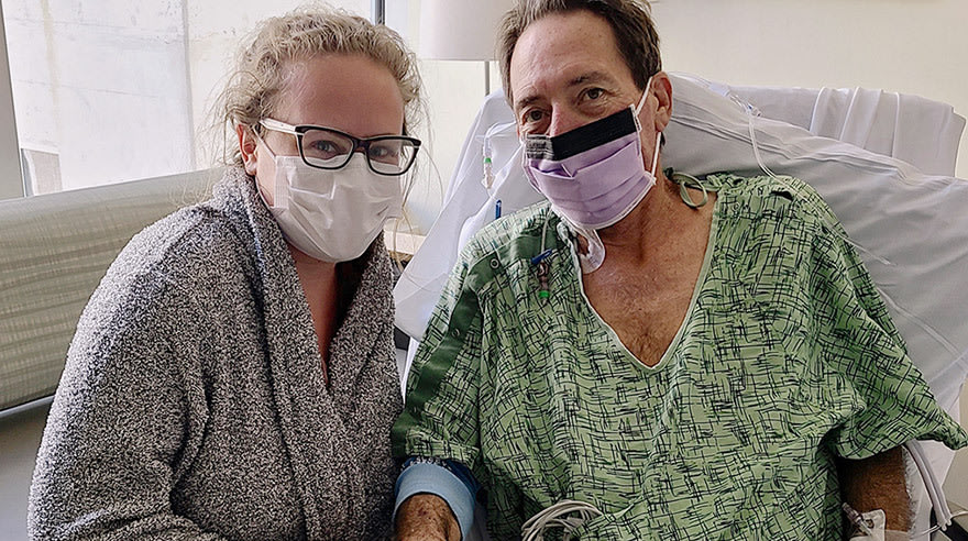 Man with face mask in hospital bed with woman with glasses and face mask by his side