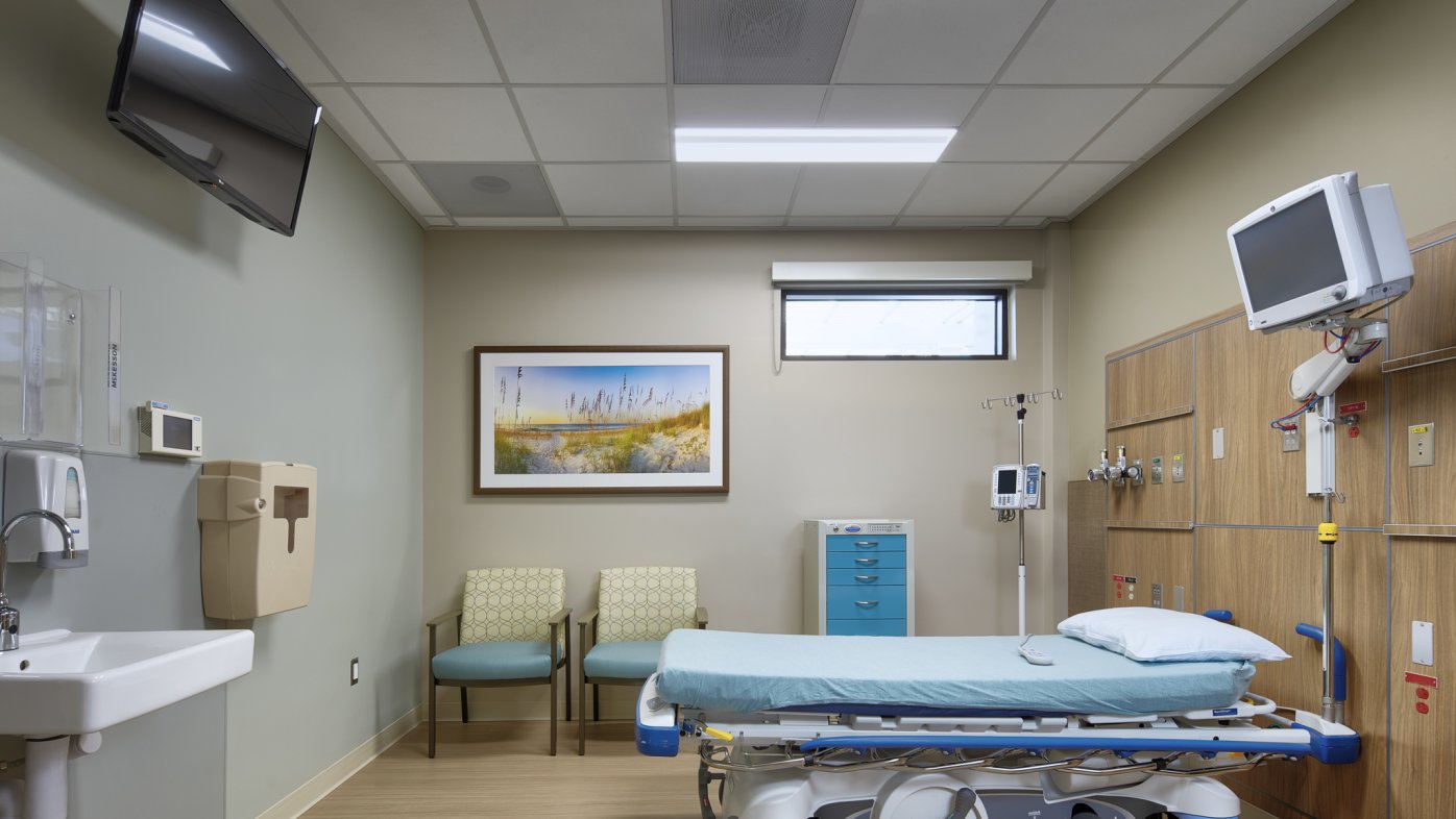 Hospital room with sink on left, painting on wall, two guest chairs and hospital bed on right