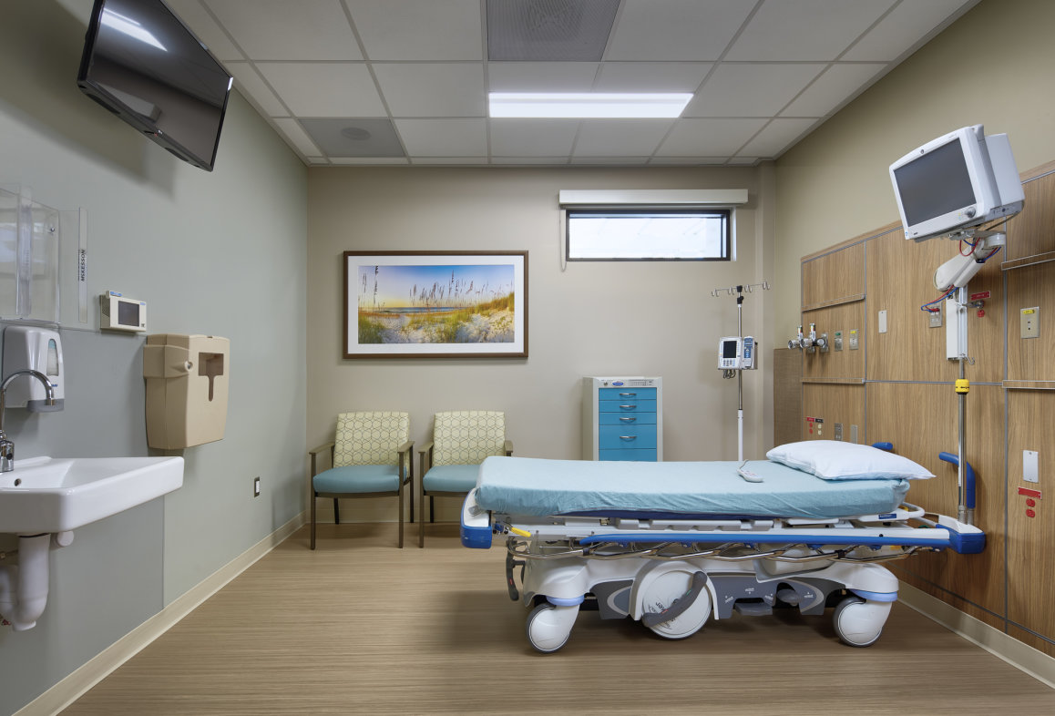 Hospital room with sink on left, painting on wall, two guest chairs and hospital bed on right
