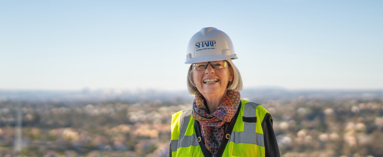 Woman smiling wearing Sharp HealthCare hardhat with city in background.
