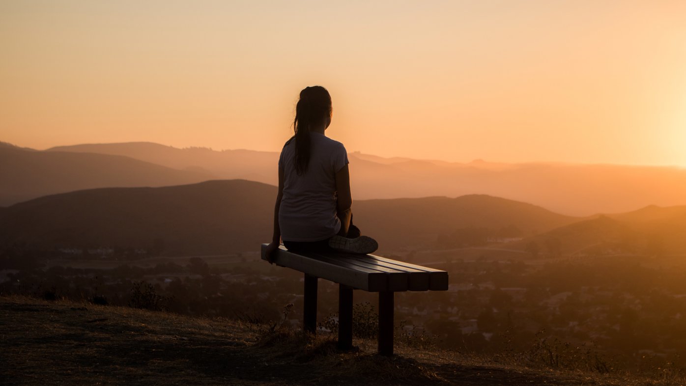 Woman sitting on a bench, watching the sunset over mountains. 