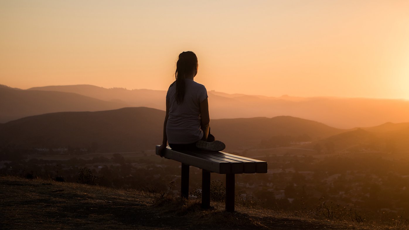 Woman sitting on a bench, watching the sunset over mountains.