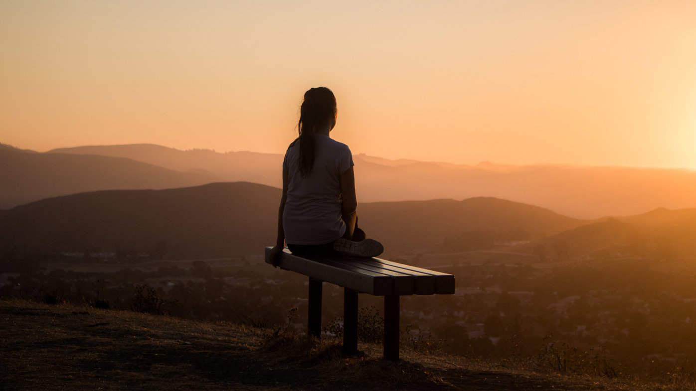 Woman sitting on a bench, watching the sunset over mountains. 