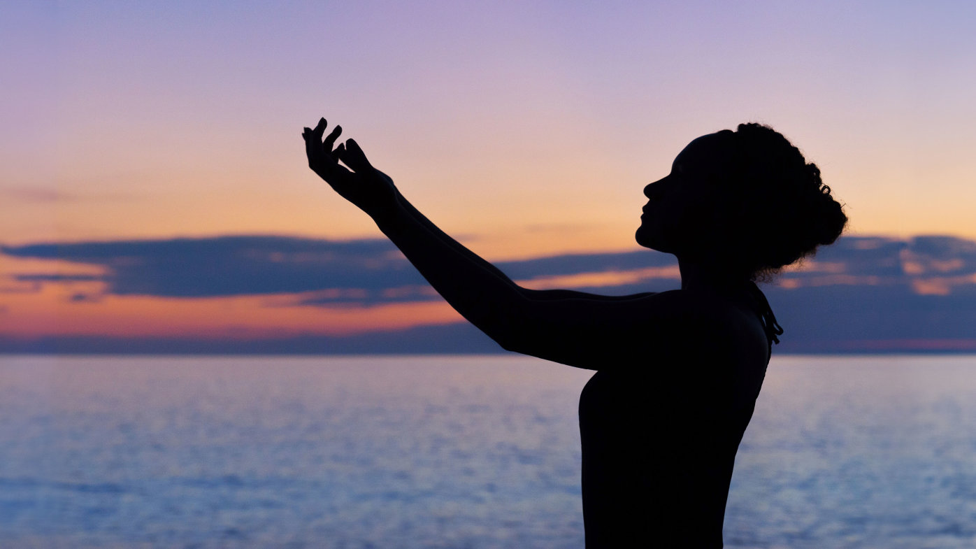 Silhouette of woman with her arms raised toward the sky in front of the ocean at sunset. 