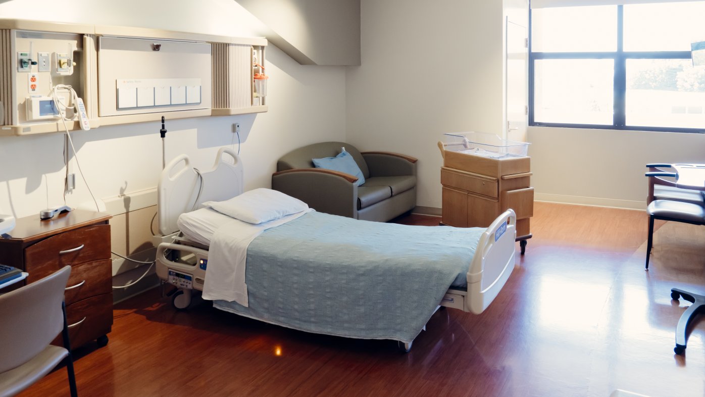 Hospital room with hospital bed, green couch, wooden floors and windows