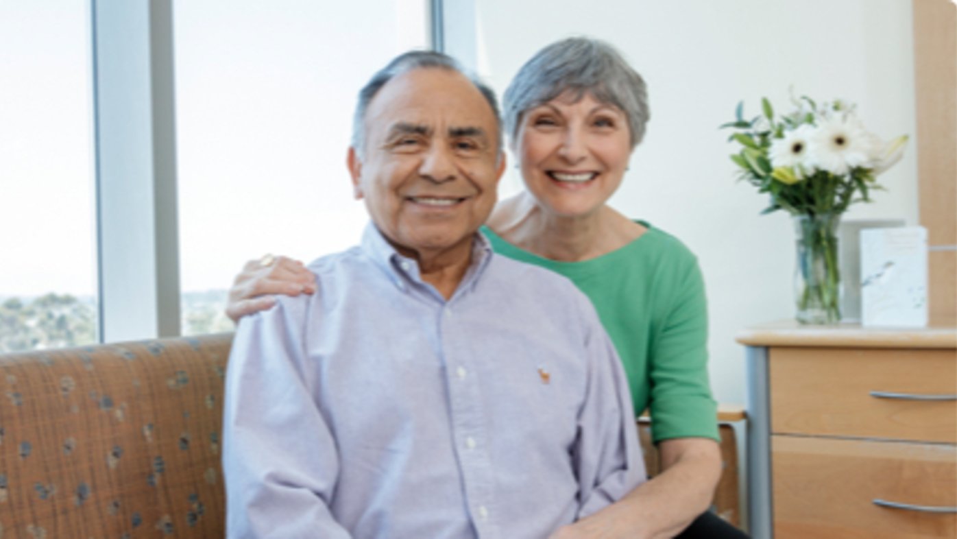 An older man in a blue button-down shirt and woman in a green sweater sit together smiling