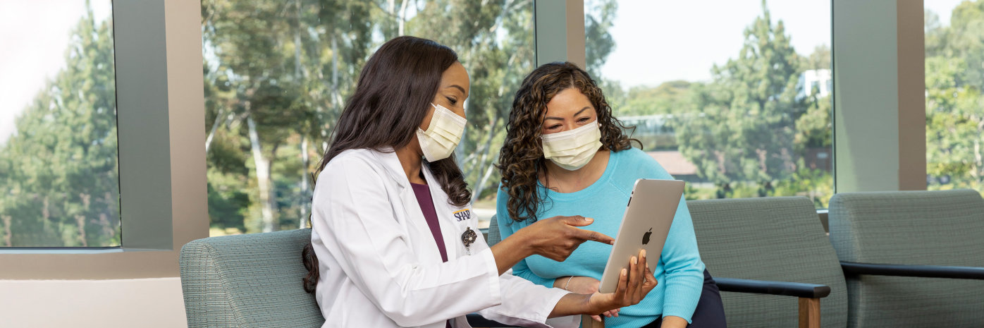 Female doctor in while lab coat and mask on left with female patient in blue shirt and mask on right looking at iPad.