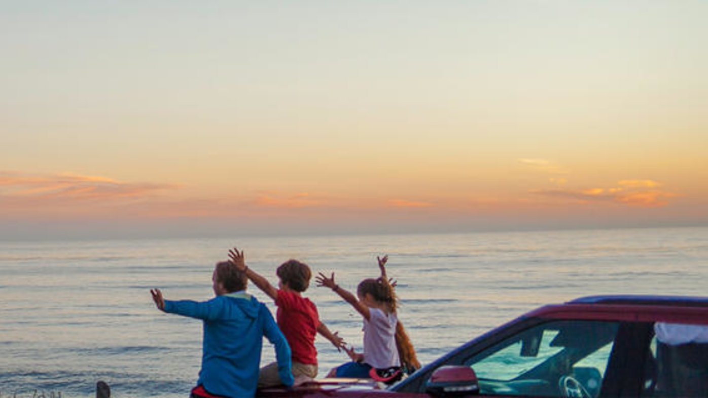 Man with children sit atop a car to watch the sunset at the beach