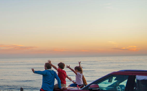 Man with children sit atop a car to watch the sunset at the beach