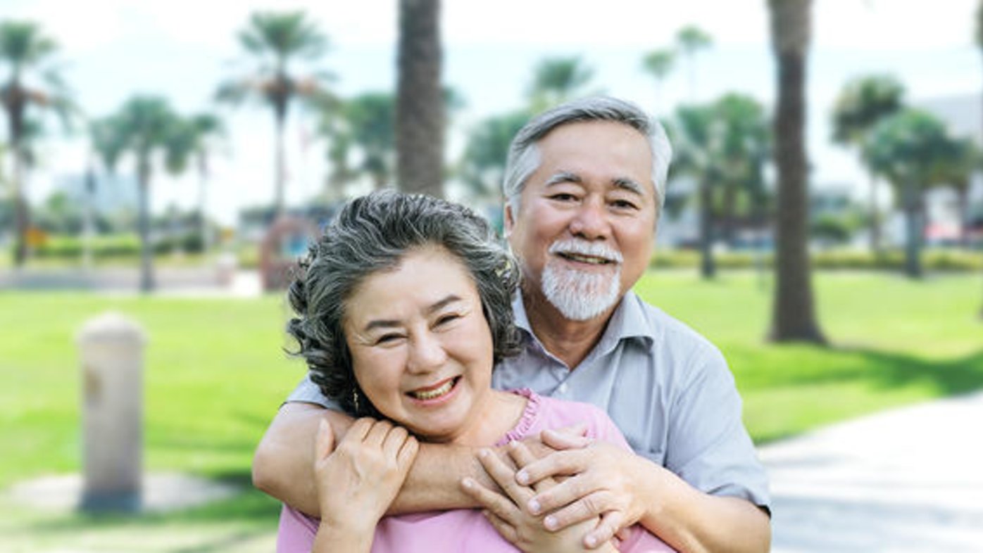 An older man and a woman in a pink shirt hug and smile outdoors at a park