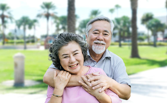 An older man and a woman in a pink shirt hug and smile outdoors at a park