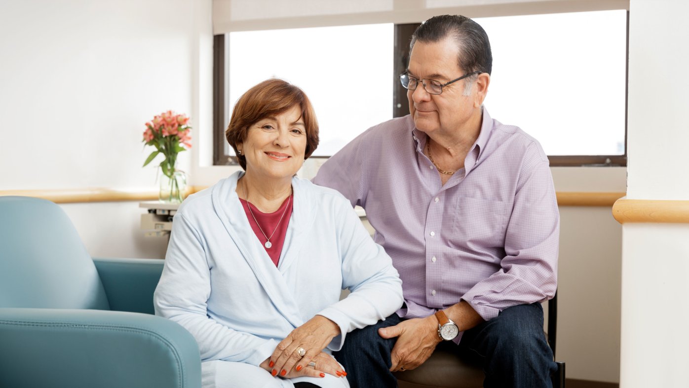 Portrait of senior couple. Woman, sitting in upholstered chair, with husband sitting slightly behind her looking at her. 