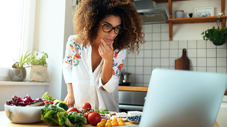 Woman standing in kitchen reading cooking recipe on laptop screen
