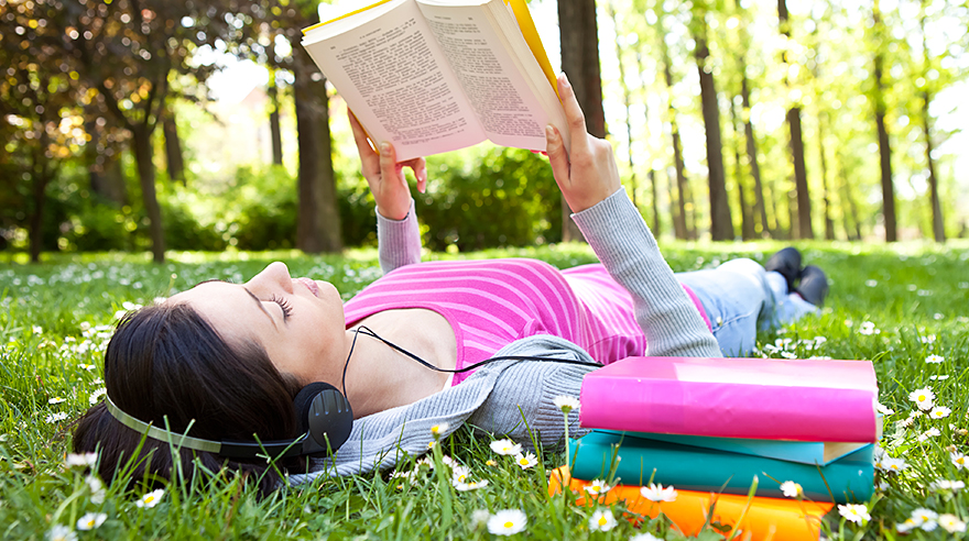 Mujer recostada al aire libre leyendo un libro y usando auriculares