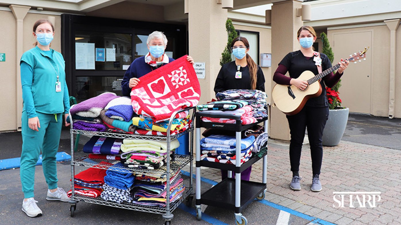 Sharp Coronado Hospital caregivers and an auxiliary volunteer stand behind stacks of handmade quilts and afghans before they were donated to patients in long-term care.
