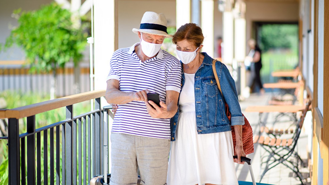 Senior couple with face masks and luggage outside on holiday.