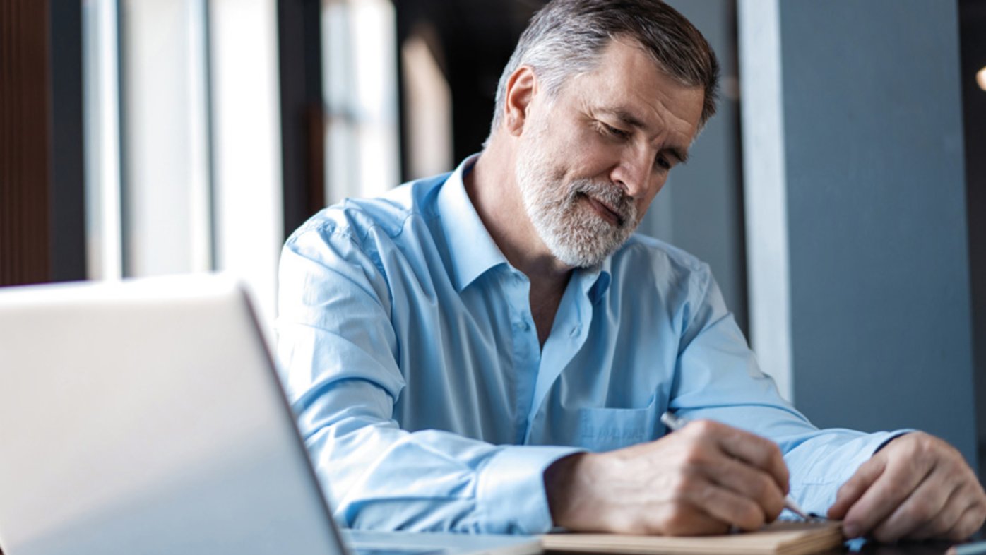 Man at desk with computer writing in notepad