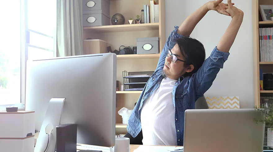 Young man stretching hands over his head while sitting at a desk in front of a computer.