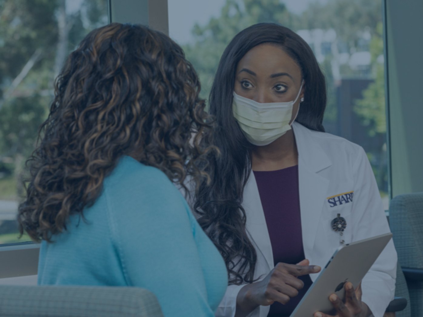 Female doctor in white lab coat on right talking to patient in blue shirt on left