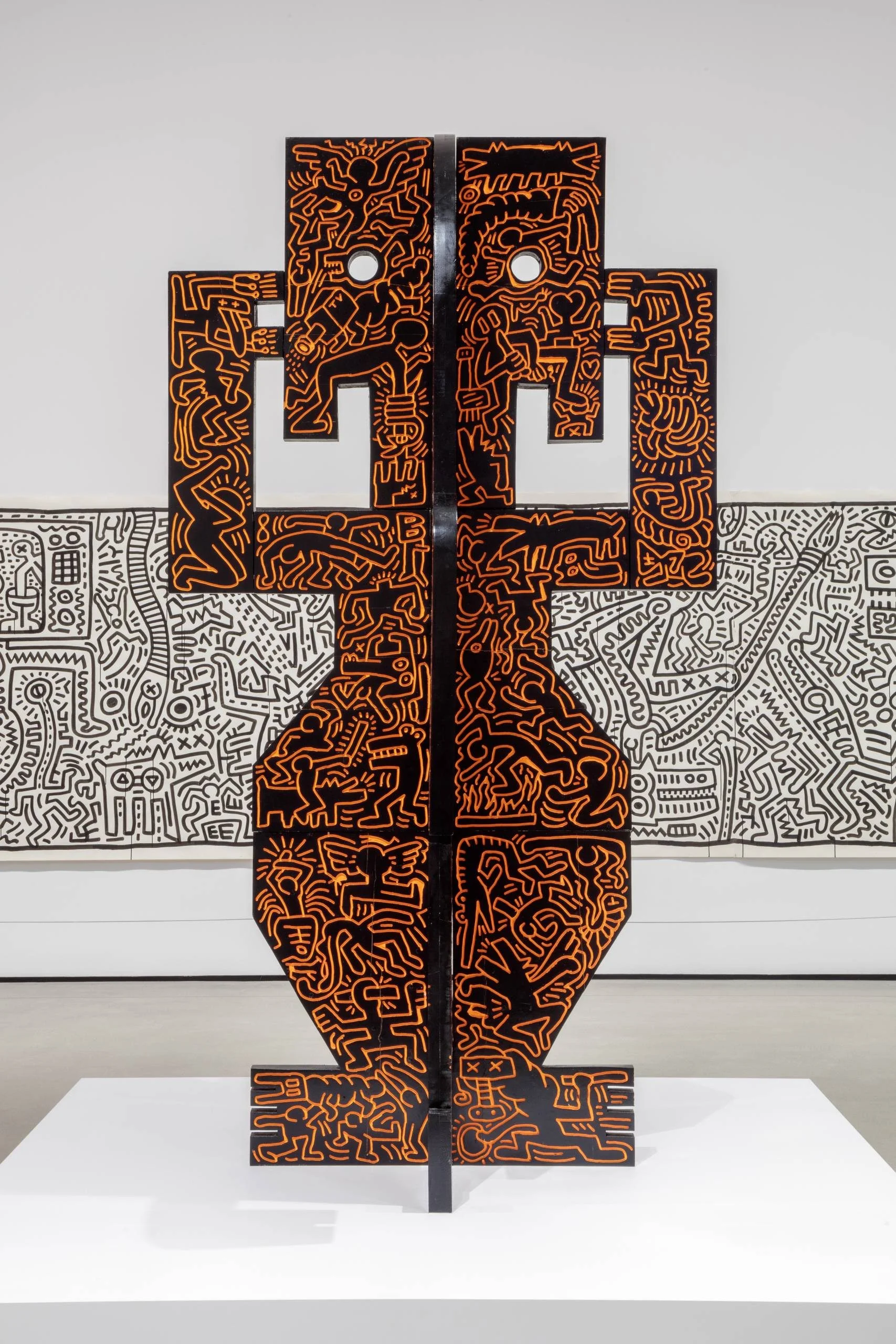 Black sculpture with orange graffiti-style figures stands before a black and white mural.
