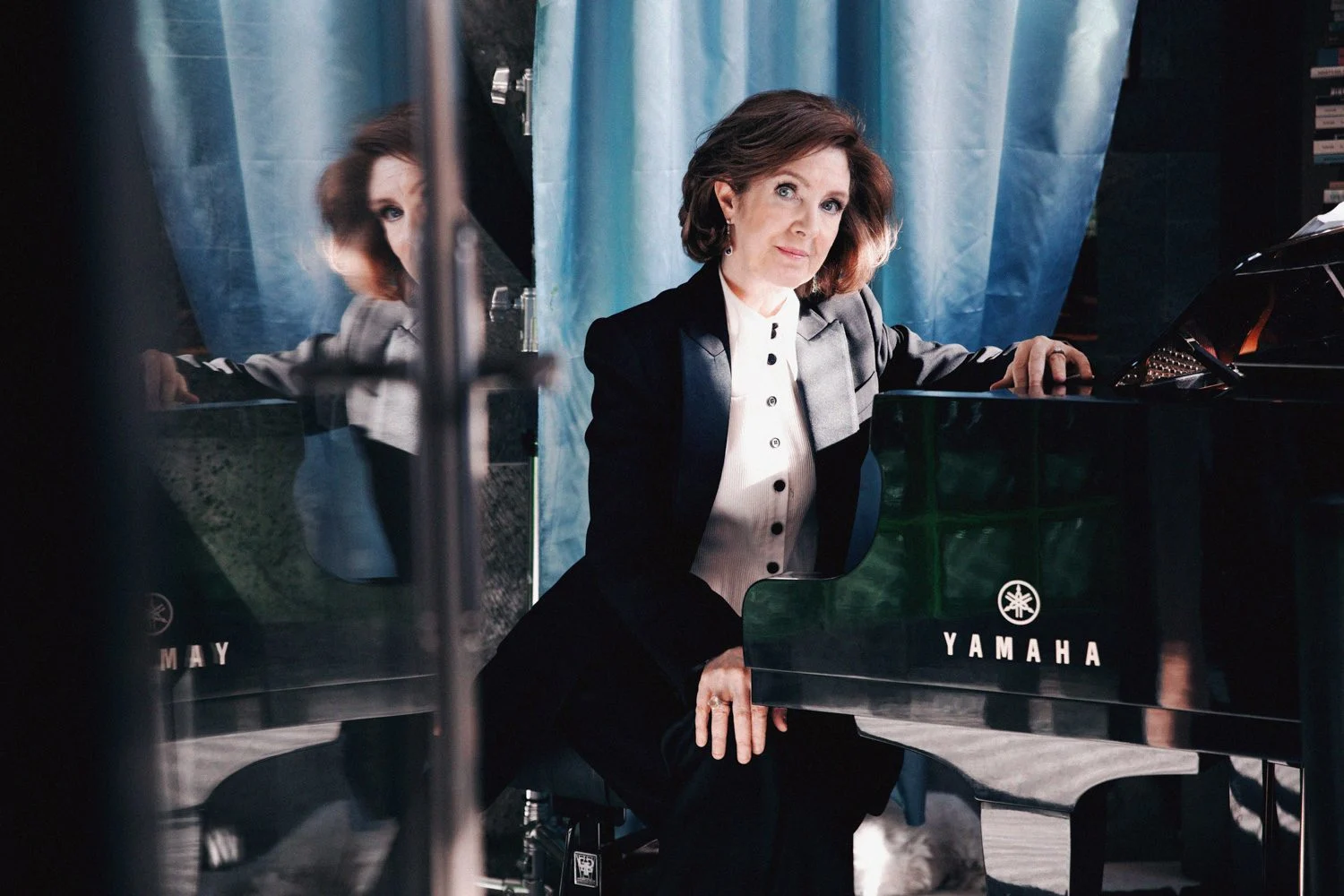 Woman in formal attire seated at a Yamaha grand piano with blue curtain background.