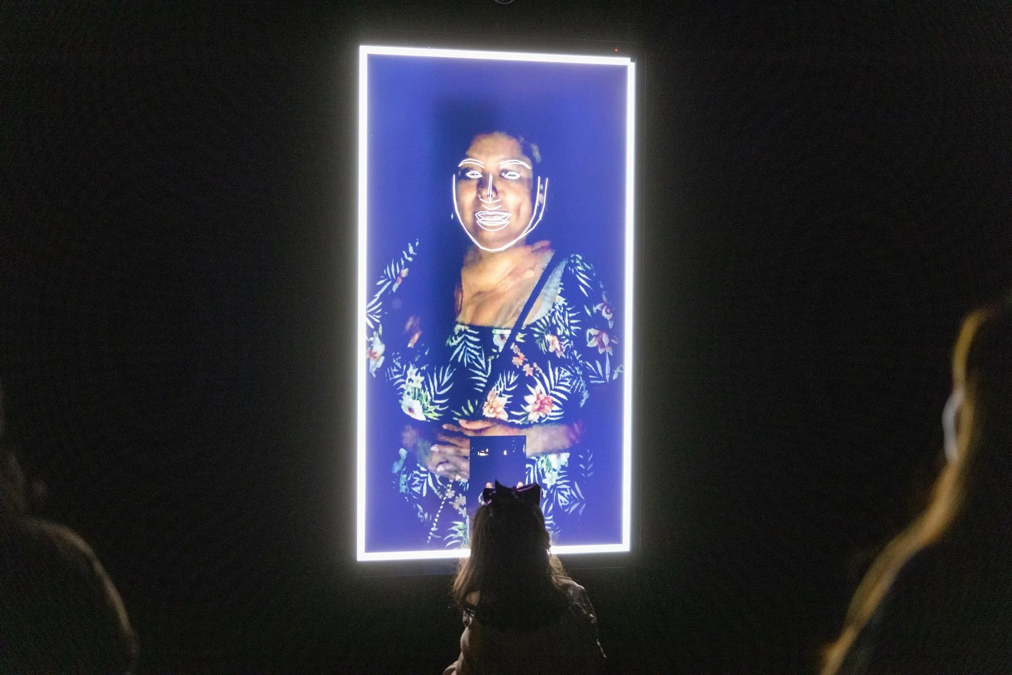 A young girl stands facing a screen outside at night that displays a face blurring and combining into a composite portrait.