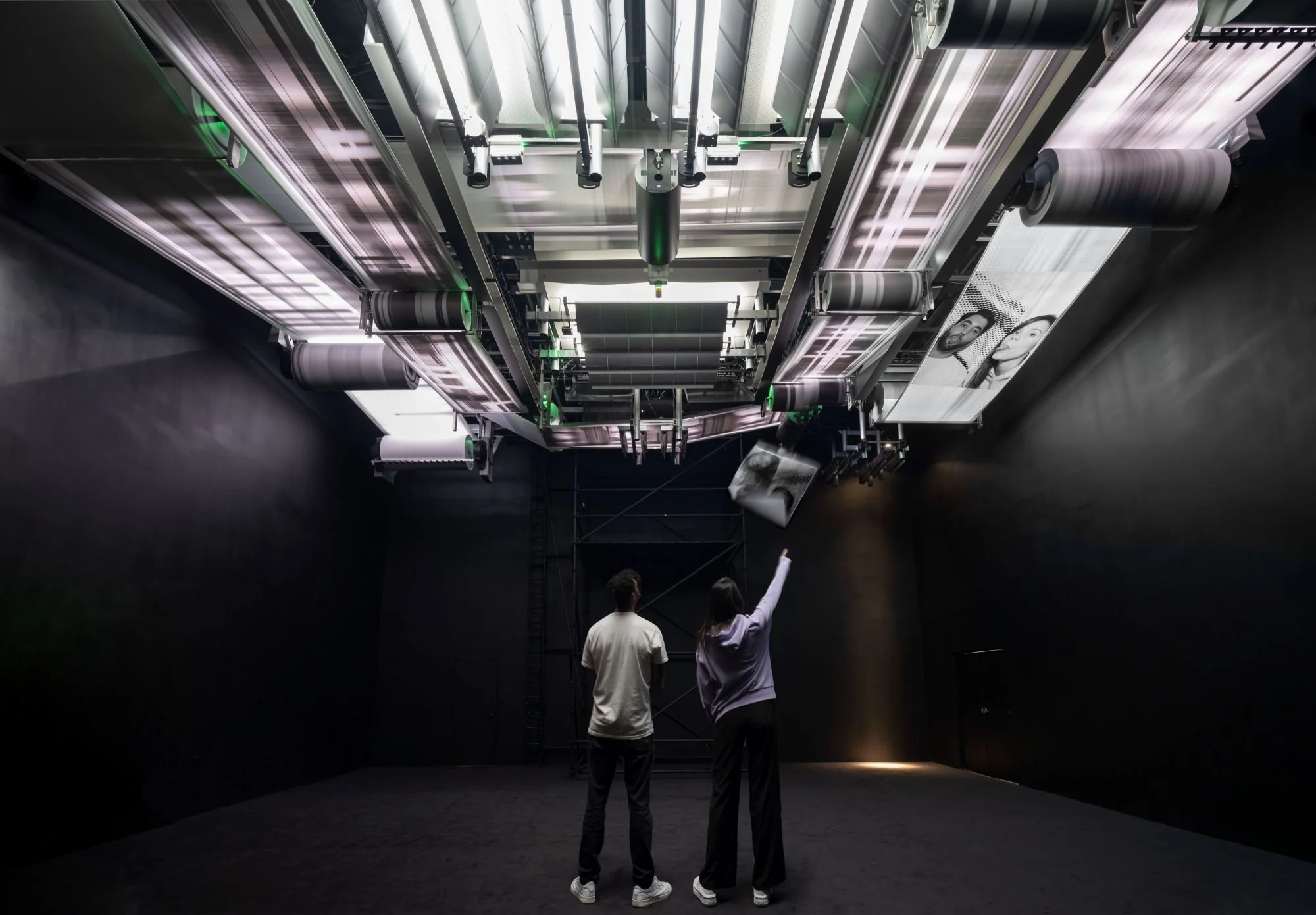 Individuals examining a ceiling-mounted printing system with rollers in a dark room.