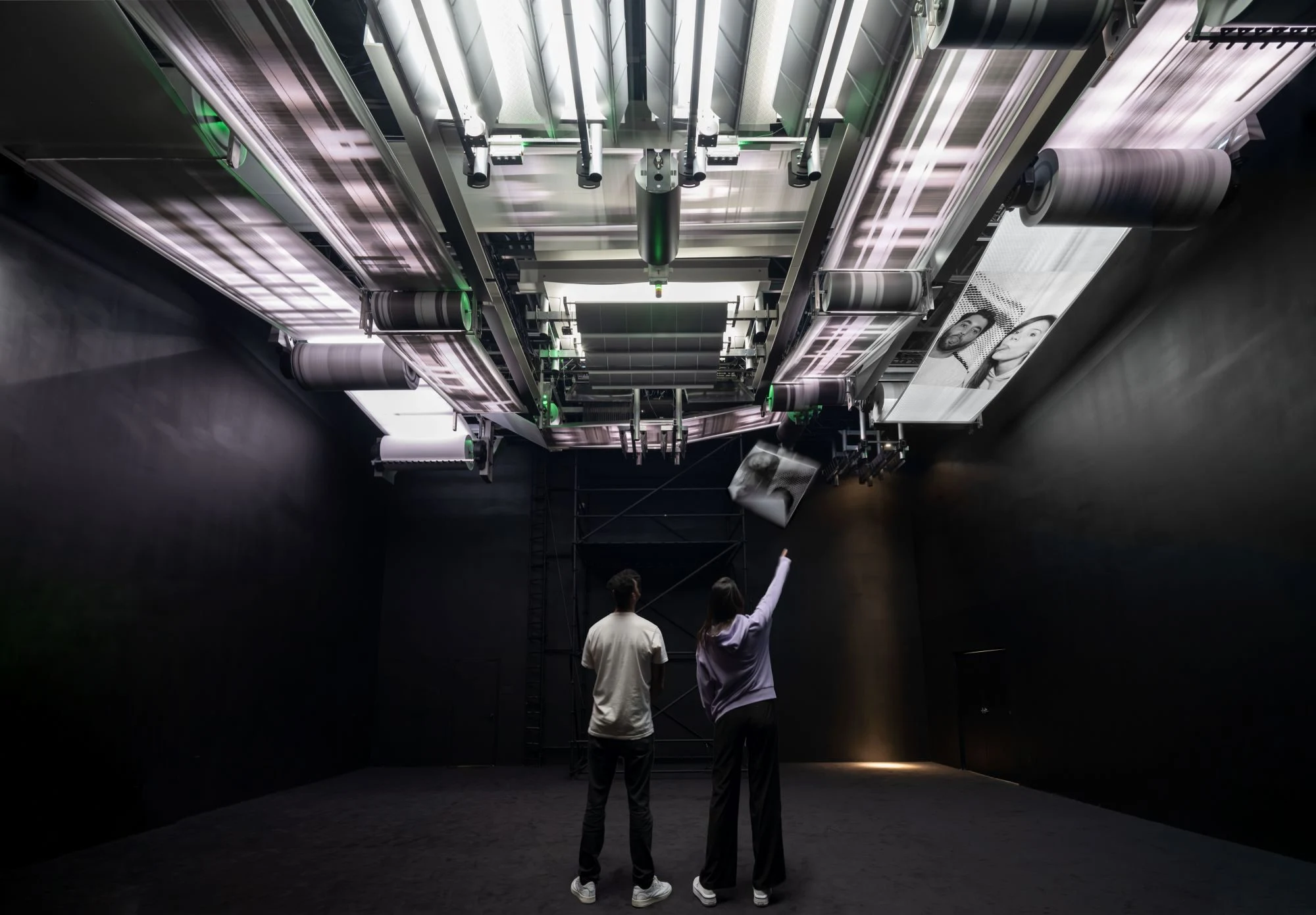 Individuals examining a ceiling-mounted printing system with rollers in a dark room.