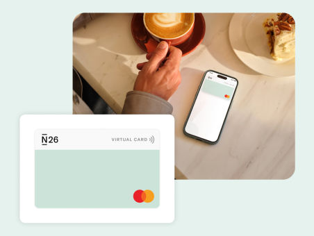 An image of an N26 virtual card with a mobile phone in the background displaying the same N26 virtual card, next to a hand holding a cappuccino cup.