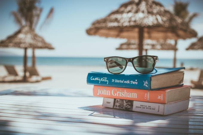 Three books and sunglasses on a table with a beach background. 