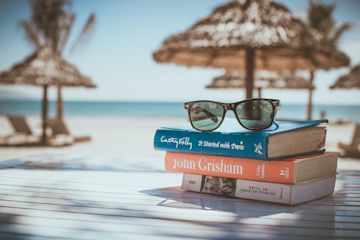 Three books and sunglasses on a table with a beach background.