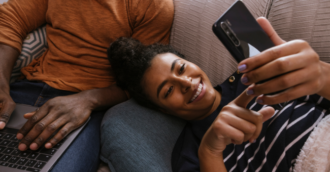 Woman lying on a sofa looking at her mobile and man working on his computer.