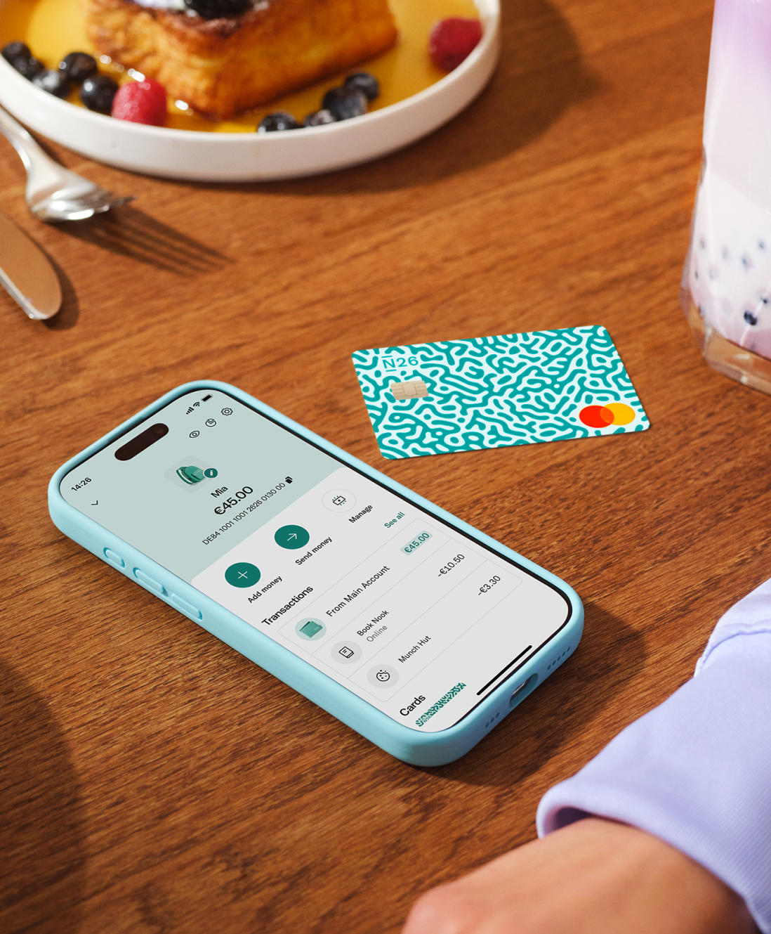 A mobile phone showing the N26 app "Under 18" interface next to a physical debit card on a wooden table with breakfast.