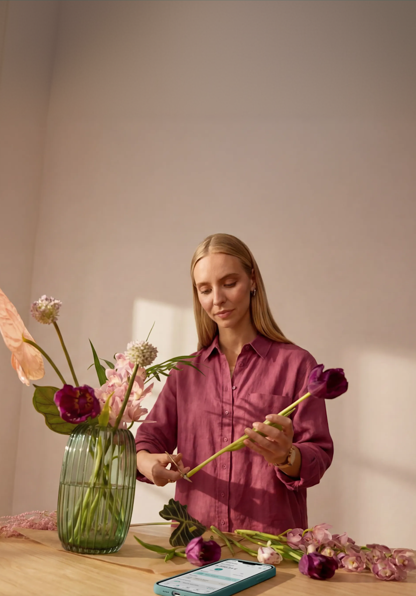 A blonde woman in a pink shirt trims fresh flower stems at a wooden table near a glass vase, with a smartphone displaying a banking app lying face-up in the foreground.