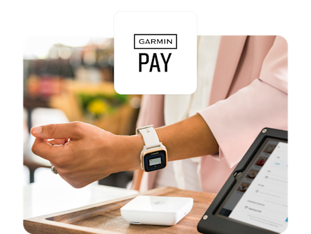 A person wearing a pink blazer uses a Garmin smartwatch to make a contactless payment at a checkout counter.