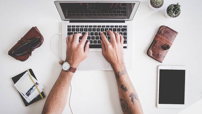 Un uomo con le braccia tatuate che scrive al computer su un tavolo bianco con occhiali da sole, portafoglio, taccuino e iPad.