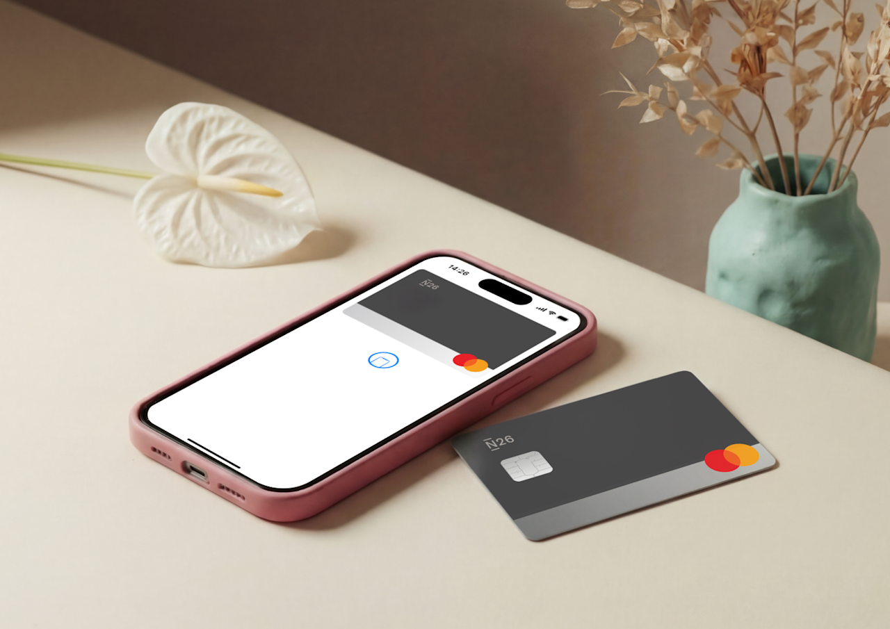 Uno smartphone rosa che mostra un portafoglio digitale e una carta fisica N26 Mastercard grigia sono appoggiati su una superficie beige accanto a piante essiccate decorative.
