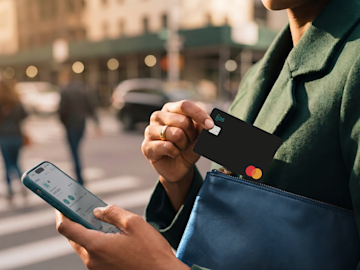 Eine Hand hält im Freien ein Smartphone, das eine digitale, pinkfarbene N26 Mastercard im Koffer-Design anzeigt.