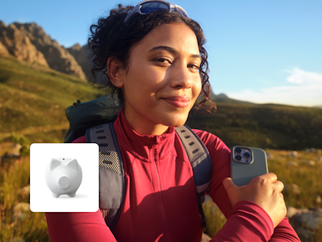 A smiling woman hiking holds a smartphone, with an icon of a white piggy bank overlaid on the image.