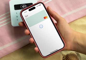 A hand holds a smartphone displaying the N26 app over a white card reader on a sandy beach.