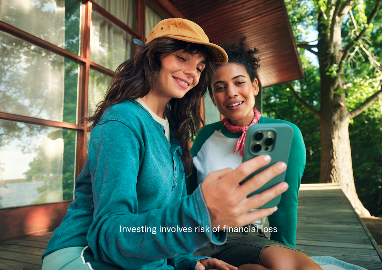Two women sit together outdoors on a wooden deck, smiling while looking at a smartphone held by one of them.