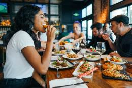 Woman in a restaurant eating a delicious piece of chicken in company of her friends. 