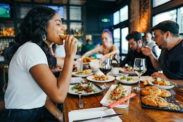 Woman in a restaurant eating a delicious piece of chicken in company of her friends.