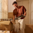 A man smiles while leaning over a box to unpack vinyl records in a bright room.