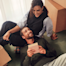 A smiling couple sits on the floor surrounded by cardboard moving boxes while looking at a small book.