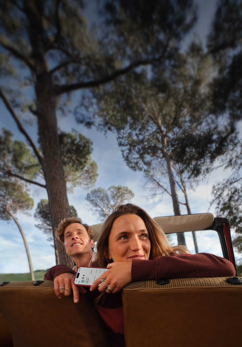 Una pareja sonriente está sentada en la parte trasera de un vehículo, mirando un paisaje natural y soleado.