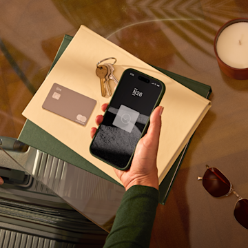 Une personne tenant un téléphone près d'une valise, avec des clés, une carte de crédit, des lunettes de soleil et une bougie sur un bureau.