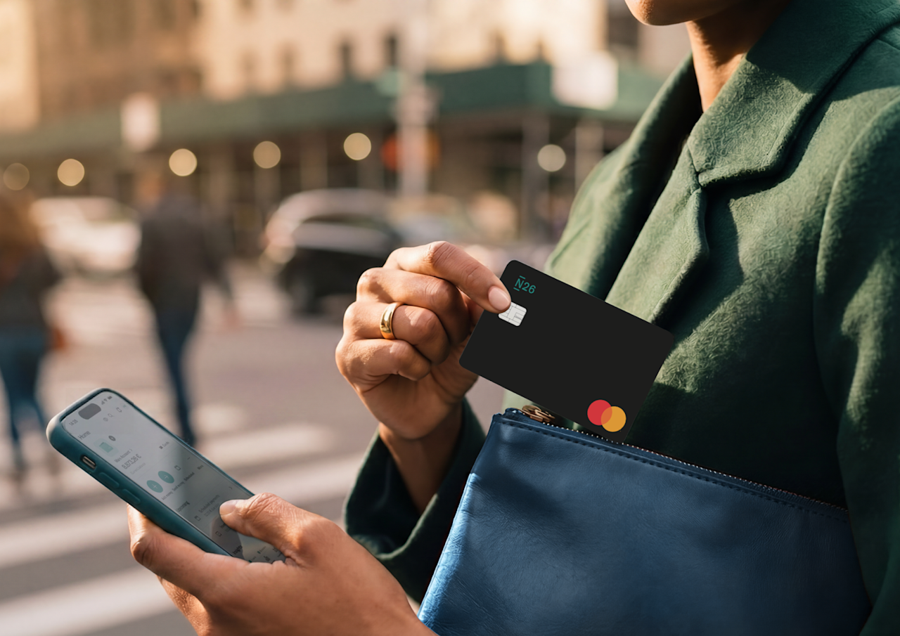 A person holds a smartphone while sliding a black N26 debit card into a blue pouch outdoors.